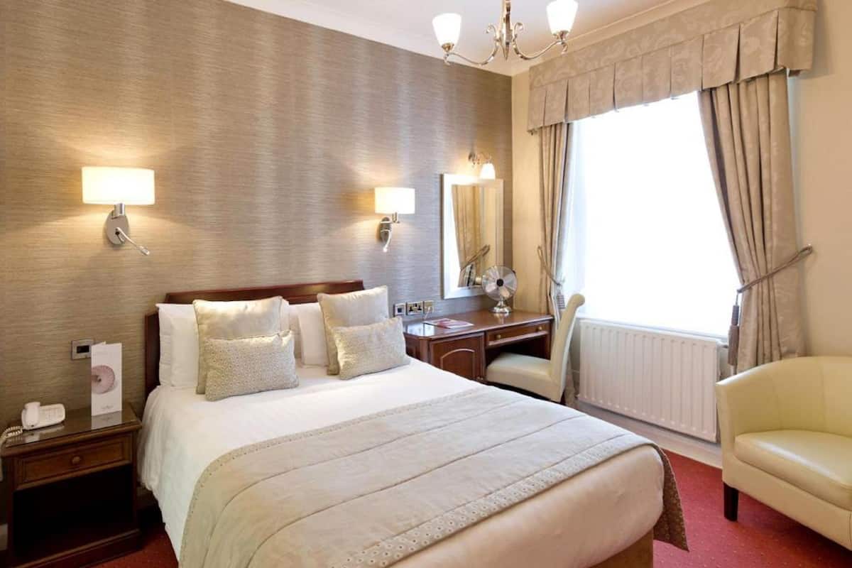 Quarto de hotel com uma cama grande, uma escrivaninha, uma cadeira e uma janela ampla com cortinas.