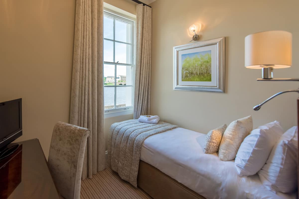 Quarto de hotel com cama, escrivaninha com televisão, cadeira, janela com cortinas, luminária na parede e quadro emoldurado.