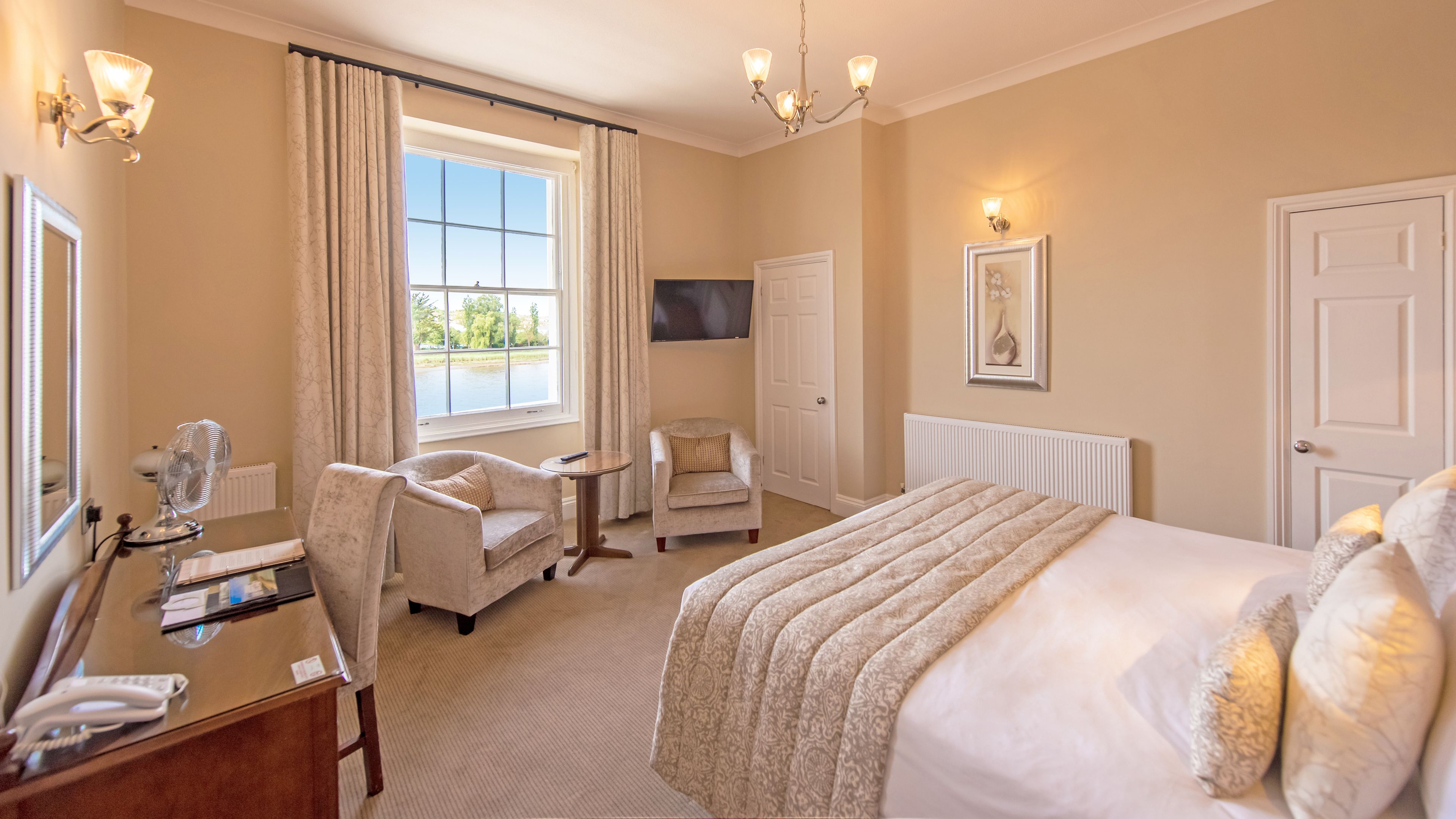 A hotel room with a large bed, a sitting area with a chair and a small table, a desk with a telephone, a television mounted on the wall, and a window with curtains.