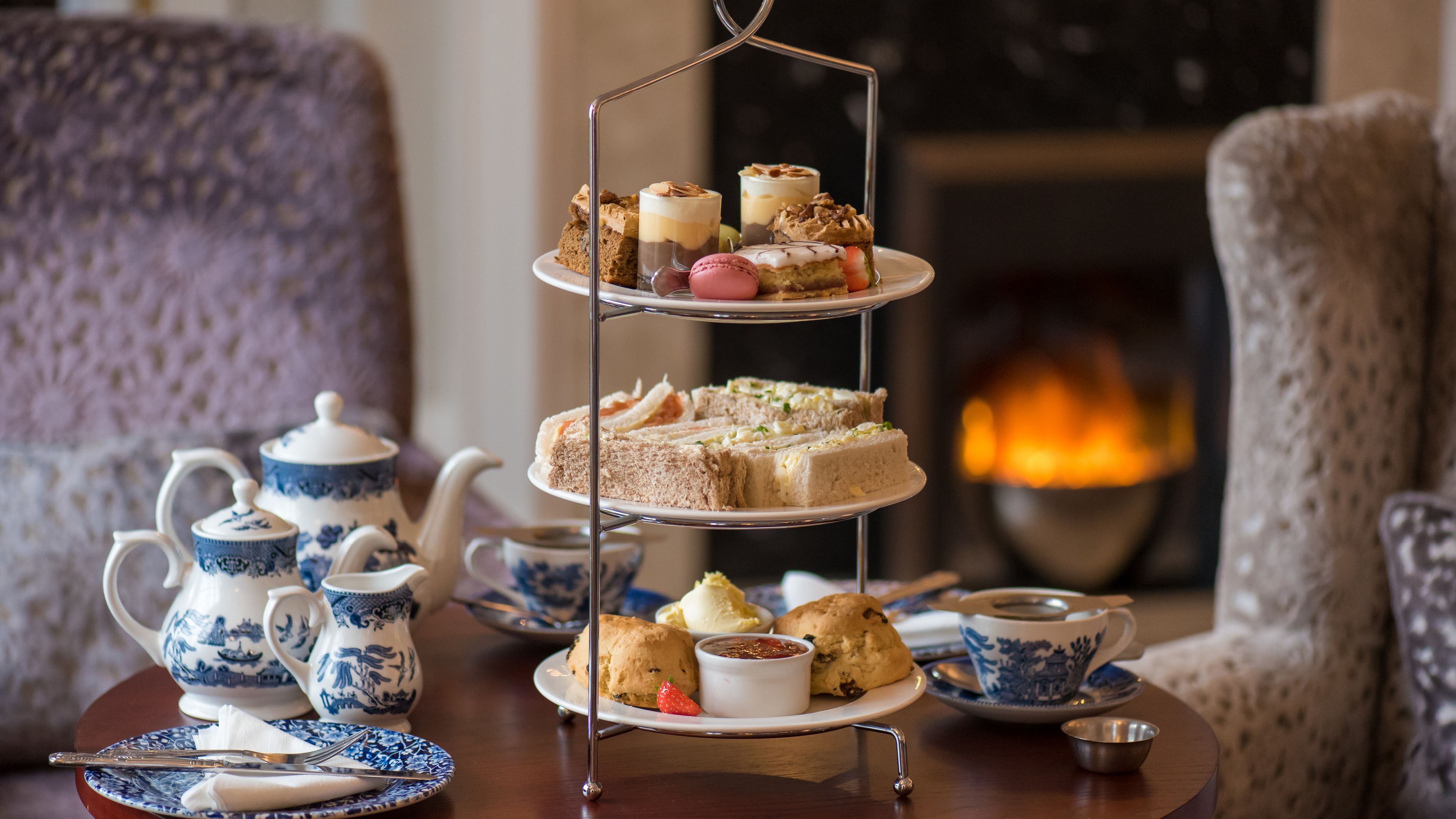 A three-tiered dessert stand with various pastries and sandwiches, surrounded by tea service and a fireplace in the background.