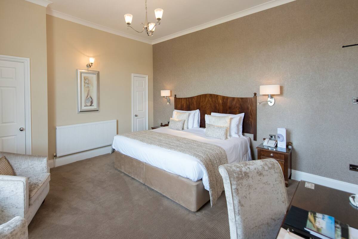 Quarto de hotel com uma cama grande, duas poltronas, uma mesa de cabeceira com abajur e uma TV fixada na parede.