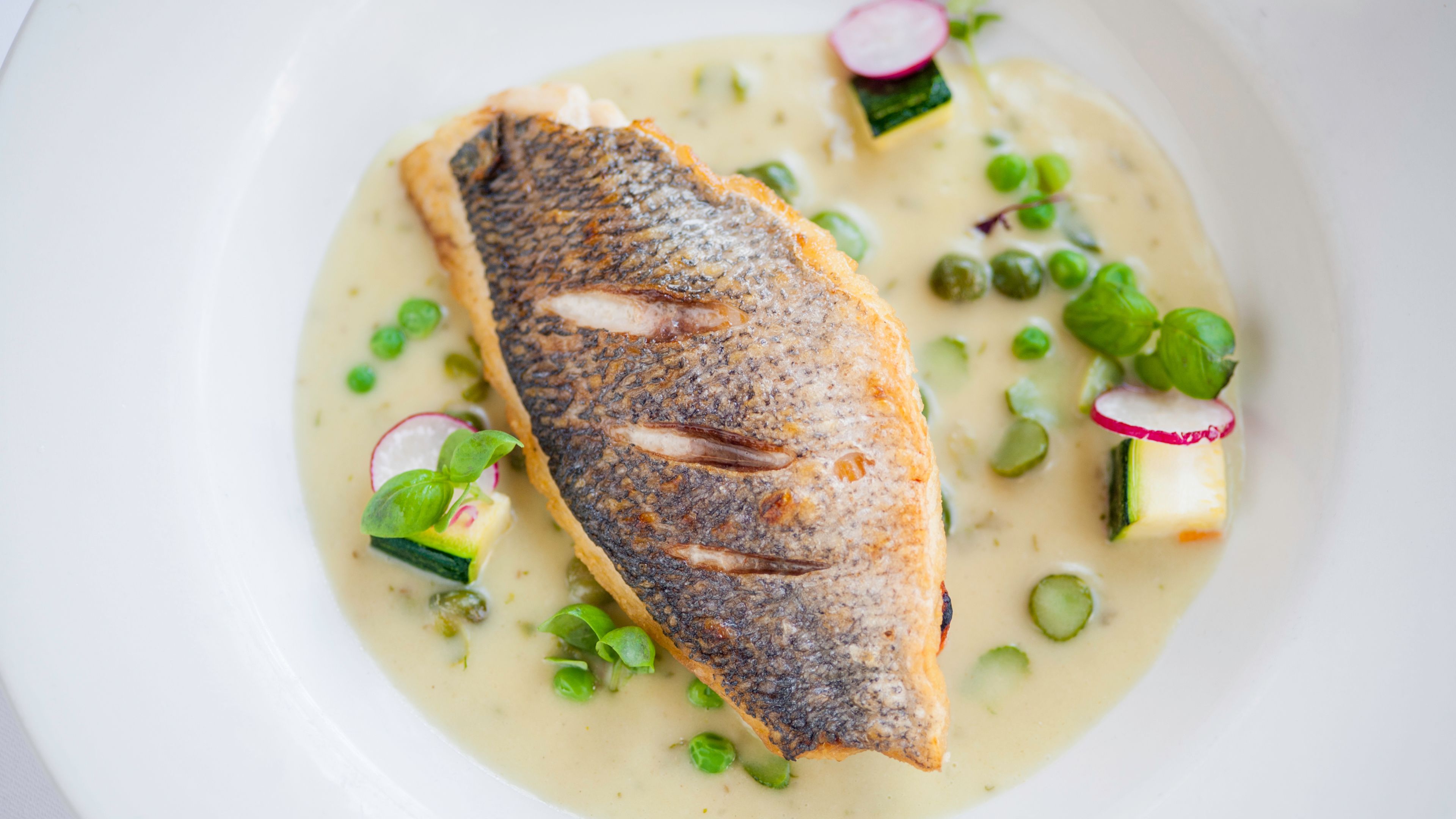 A white plate with a grilled fish, green peas, radish slices, and zucchini.