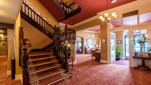 Una gran escalera con barandillas ornamentales y un suelo alfombrado en rojo que conduce a una habitación con un ventanal y una araña.