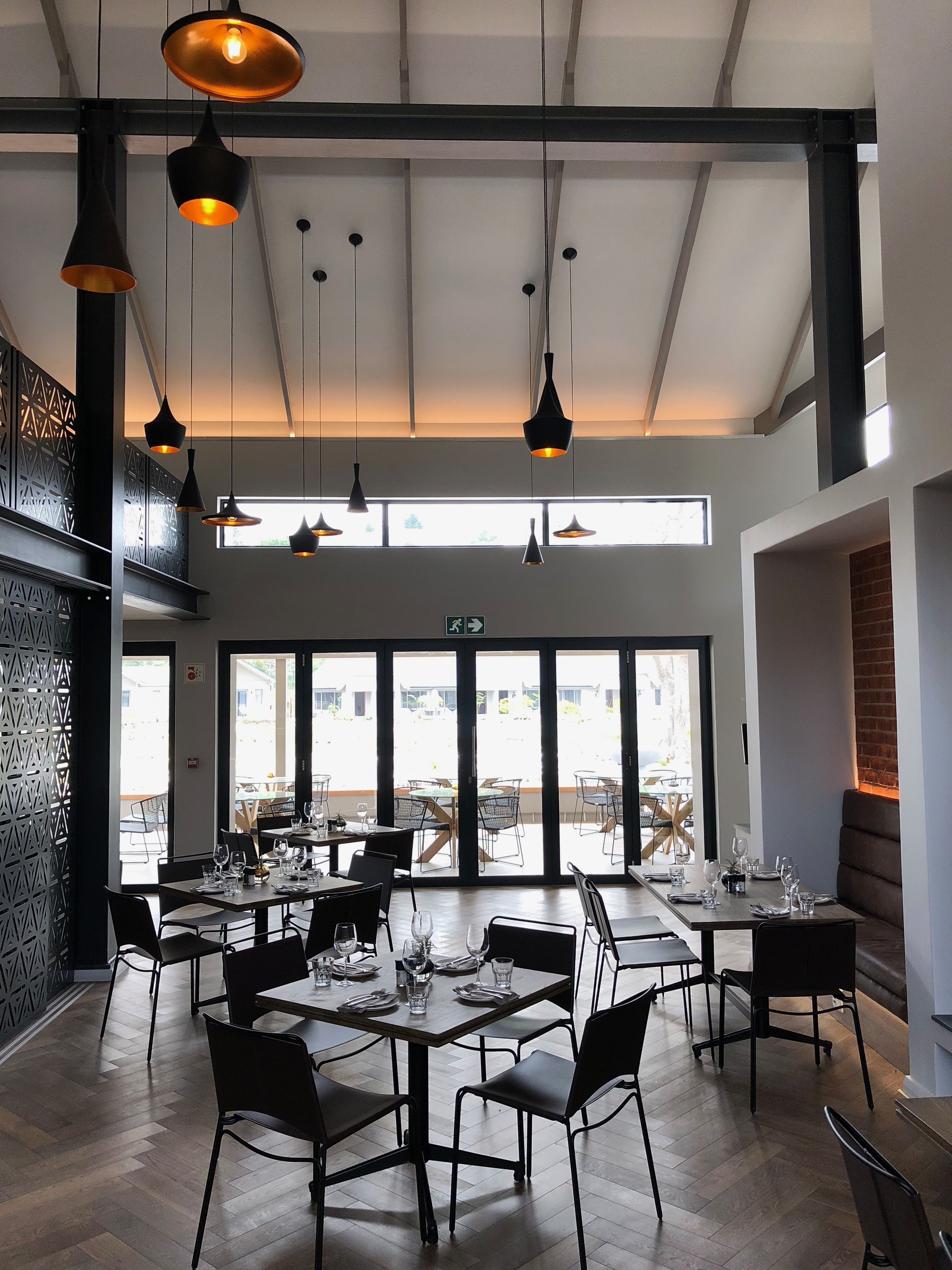 Een modern restaurant met meerdere eettafels, zwarte stoelen en hanglampen. Grote ramen zorgen voor natuurlijk licht en uitzicht op buiten.