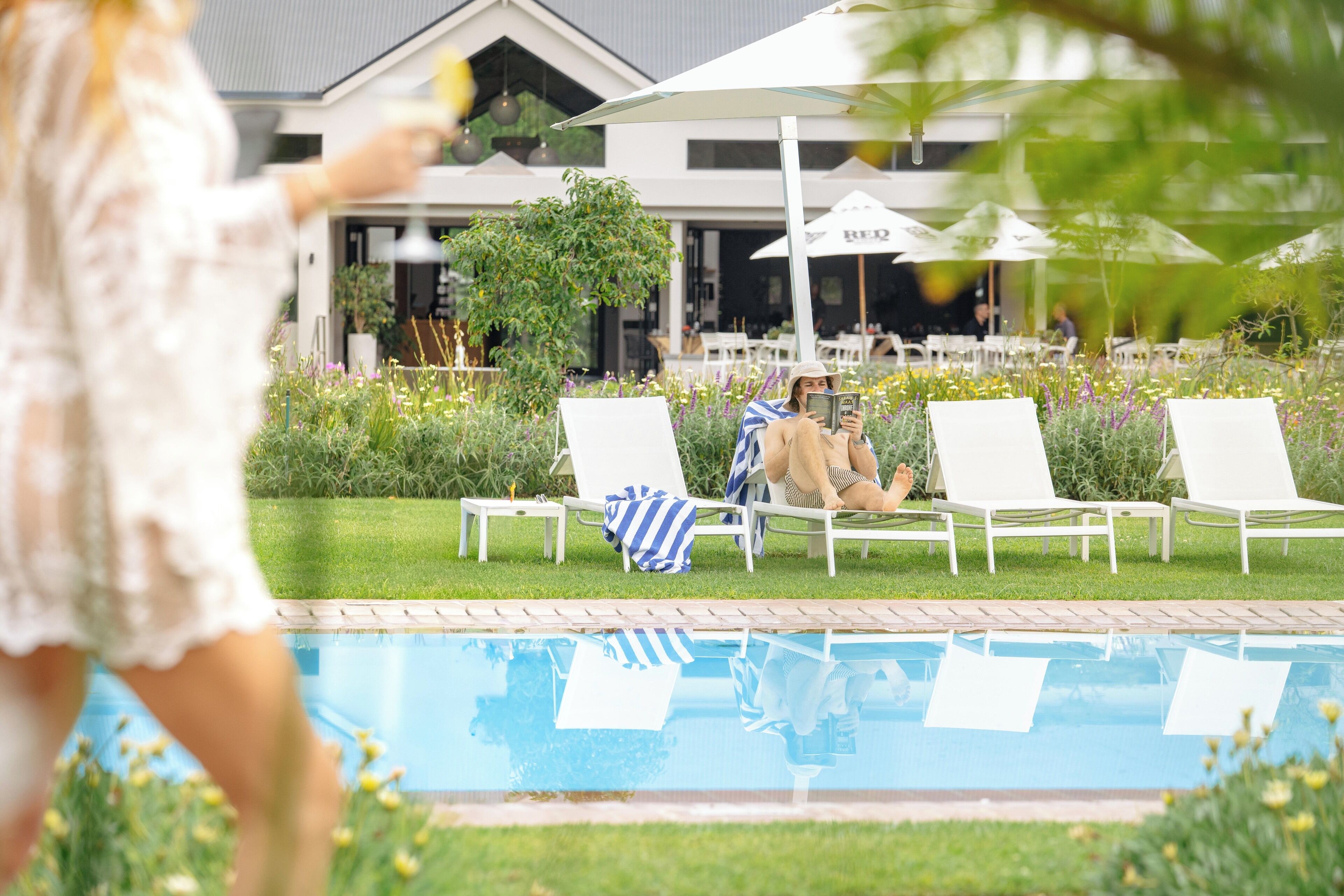 Zona de piscina amb gandules i ombrel·les, envoltada de vegetació i un edifici al fons.