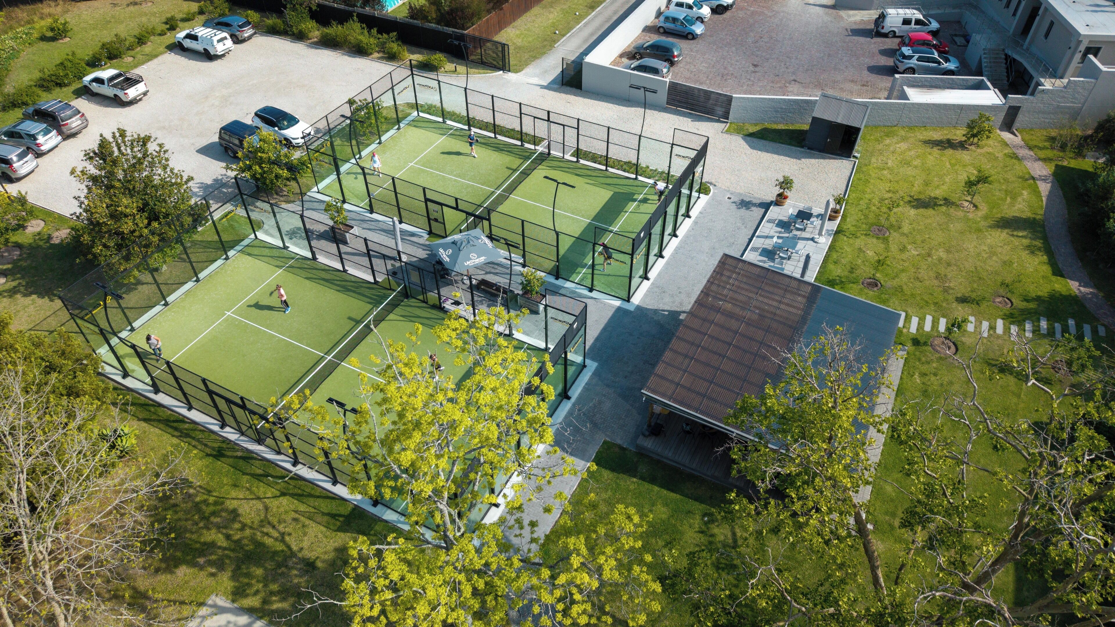 An aerial view of a tennis court complex with multiple courts, a parking area, and a building with a sloped roof.