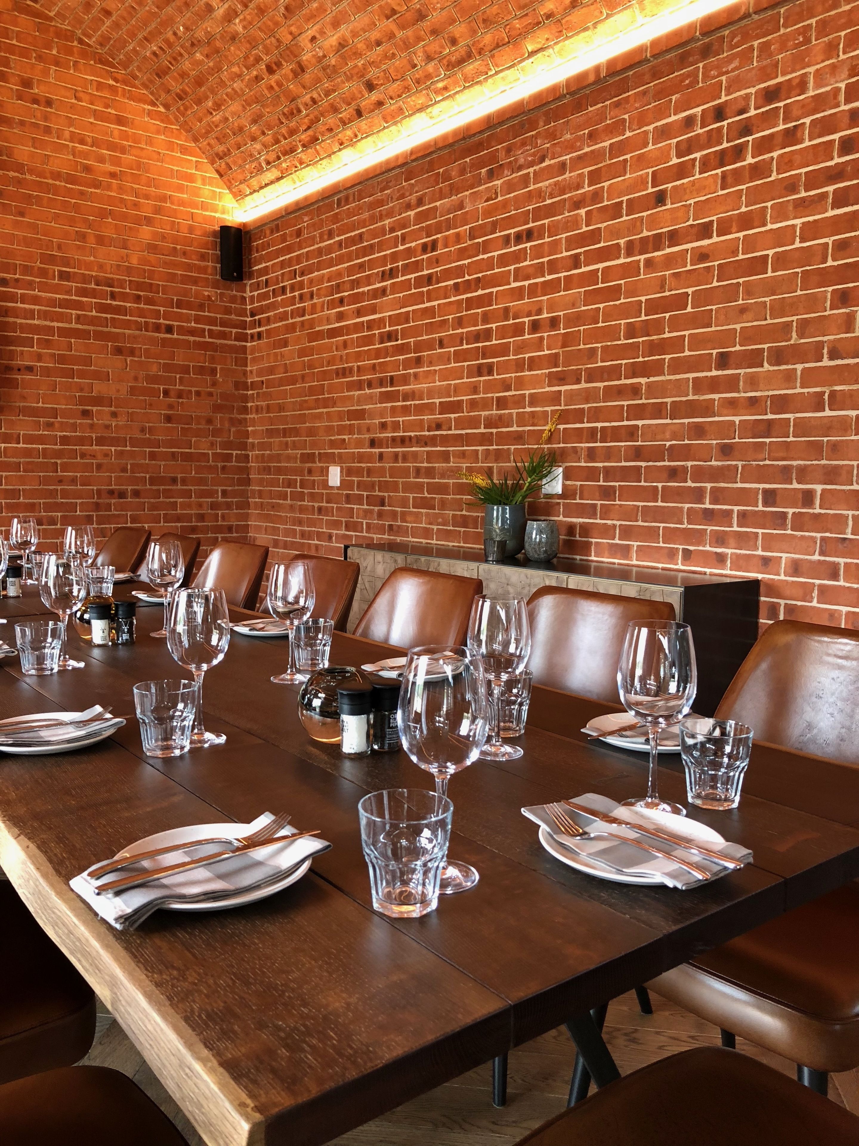 A long dining table set with wine glasses, plates, and cutlery, surrounded by leather chairs, in a room with brick walls and a wooden ceiling.