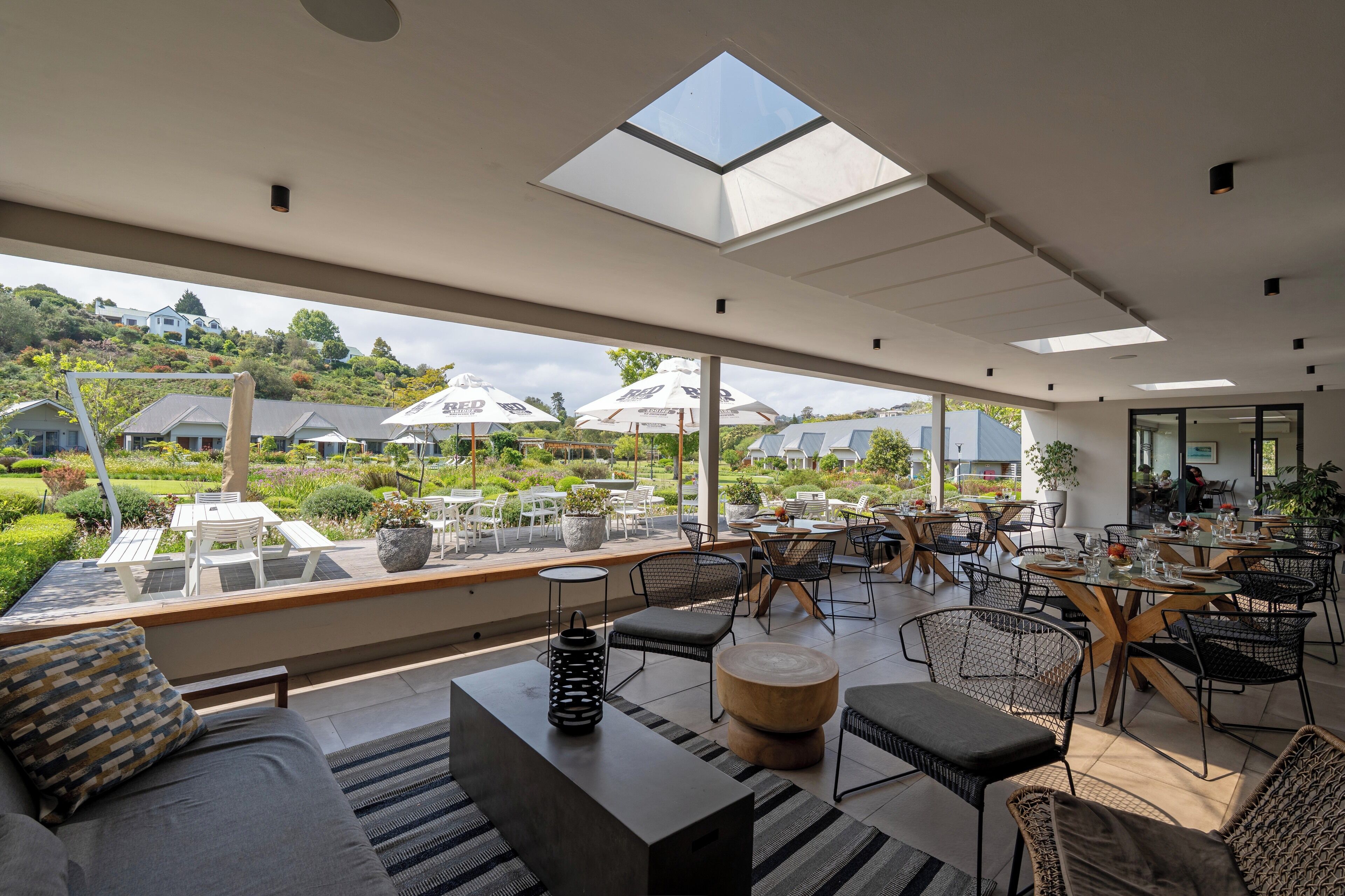 A spacious outdoor dining area with multiple tables and chairs, a large skylight, and a view of a lush garden and houses in the background.