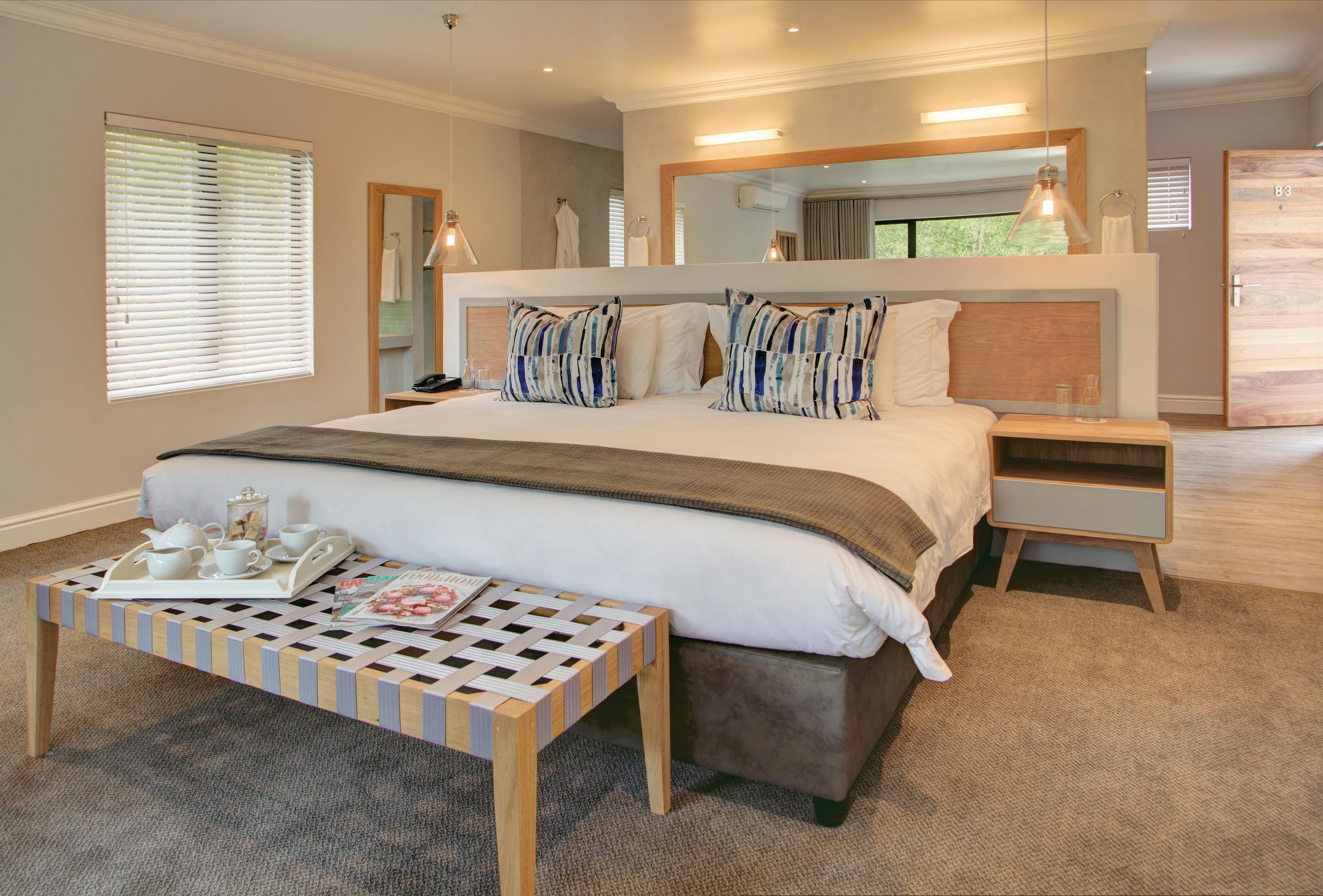 Une chambre à coucher moderne avec un grand lit, des tables de chevet, une lampe de chevet, un banc en bois avec un plateau, et un grand miroir au-dessus du lit.