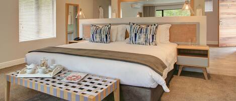 Une chambre à coucher moderne avec un grand lit, des tables de chevet, une lampe de chevet, un banc en bois avec un plateau, et un grand miroir au-dessus du lit.