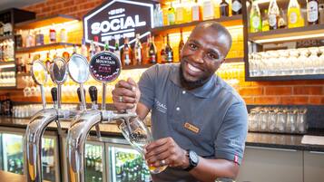 A bartender pouring a drink from a tap labeled "BLACK MOUNTAIN PINK" at a bar with shelves of bottles and glasses in the background.