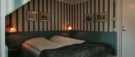 A room with two beds, striped wallpaper, framed pictures on the wall, a door, and a lamp on the left.