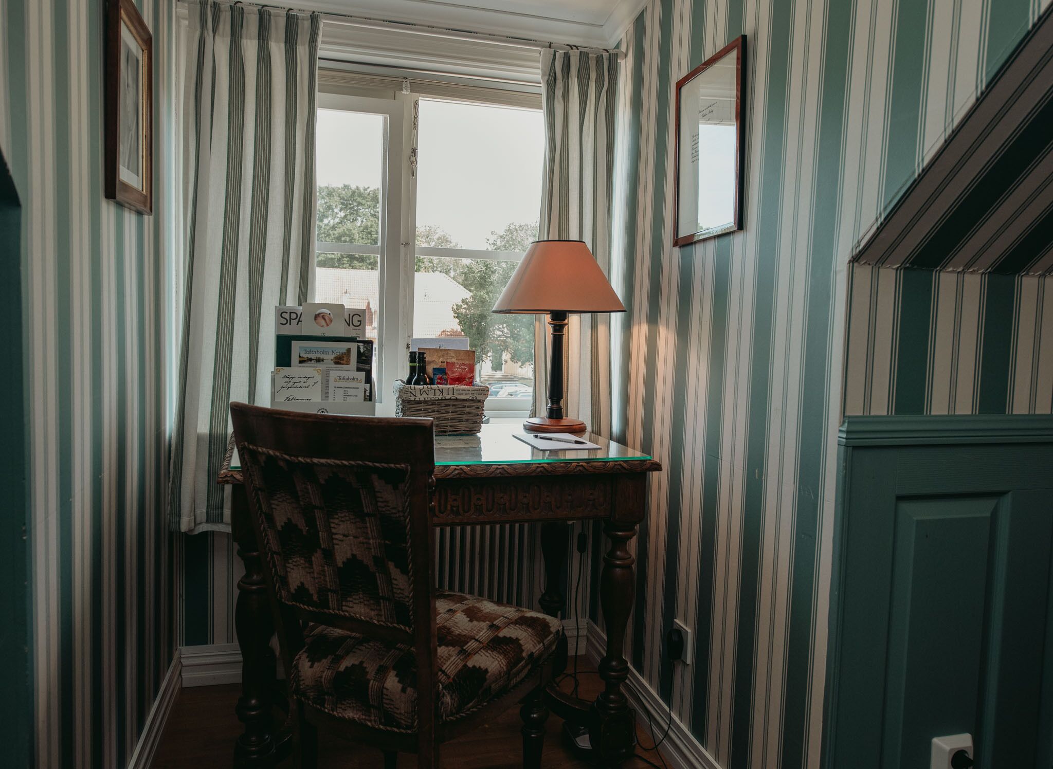 A corner room with striped wallpaper, a wooden chair, a table with a lamp, and a window with curtains.