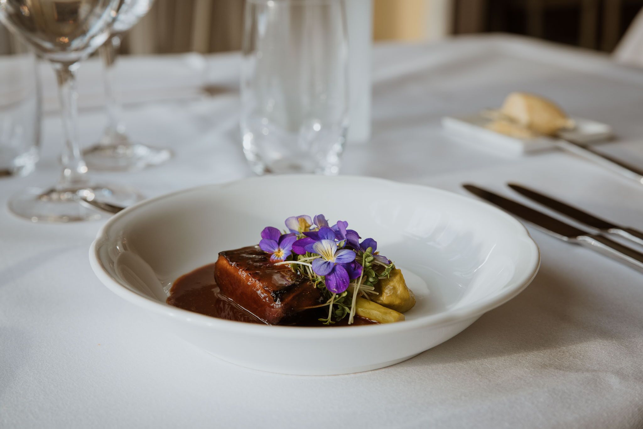A white plate with a piece of meat, purple flowers, and green garnish on a white tablecloth.