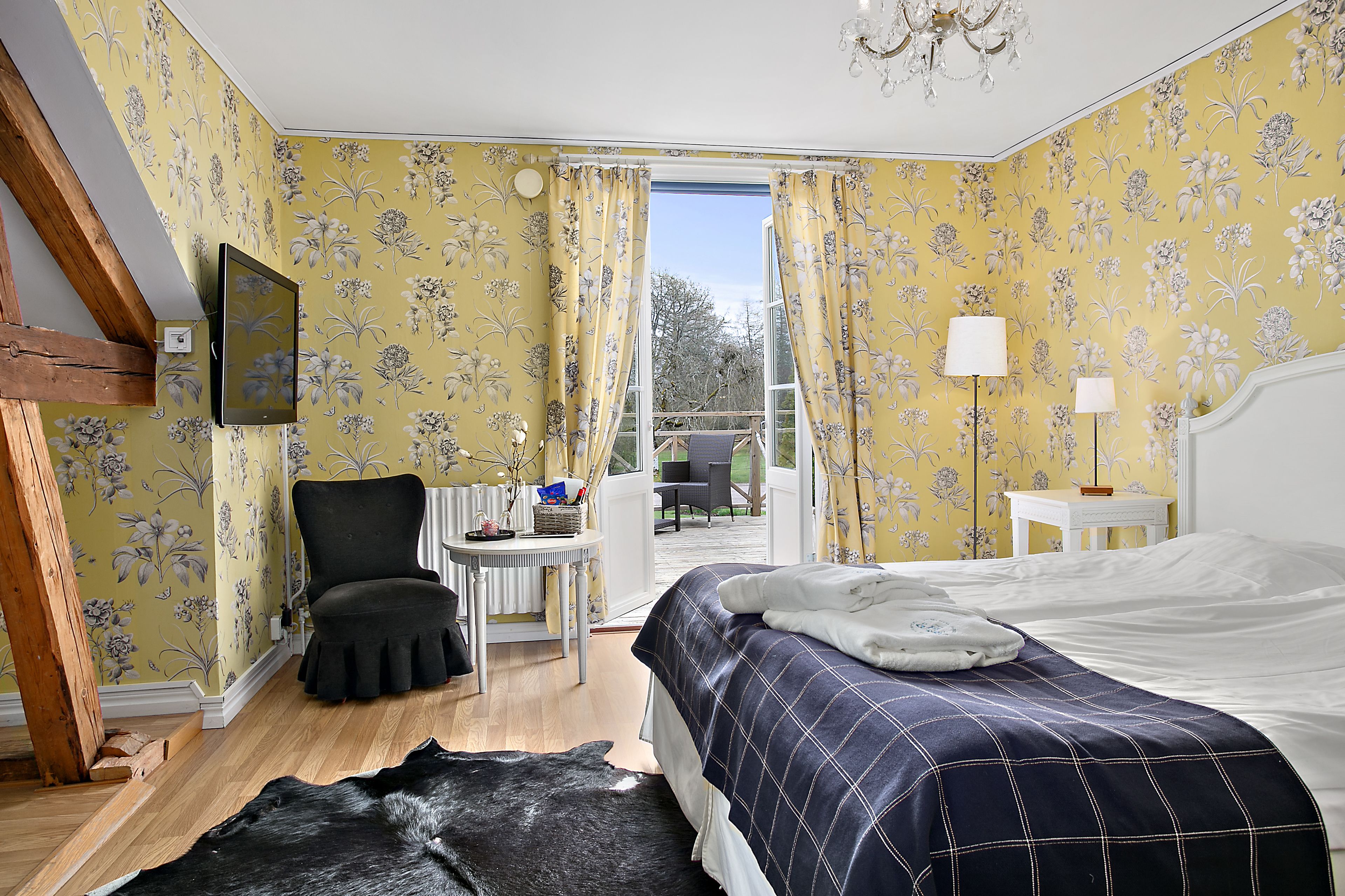 A bedroom with floral wallpaper, a bed with a checkered duvet, a chair, a small table, and a view of the outdoors.