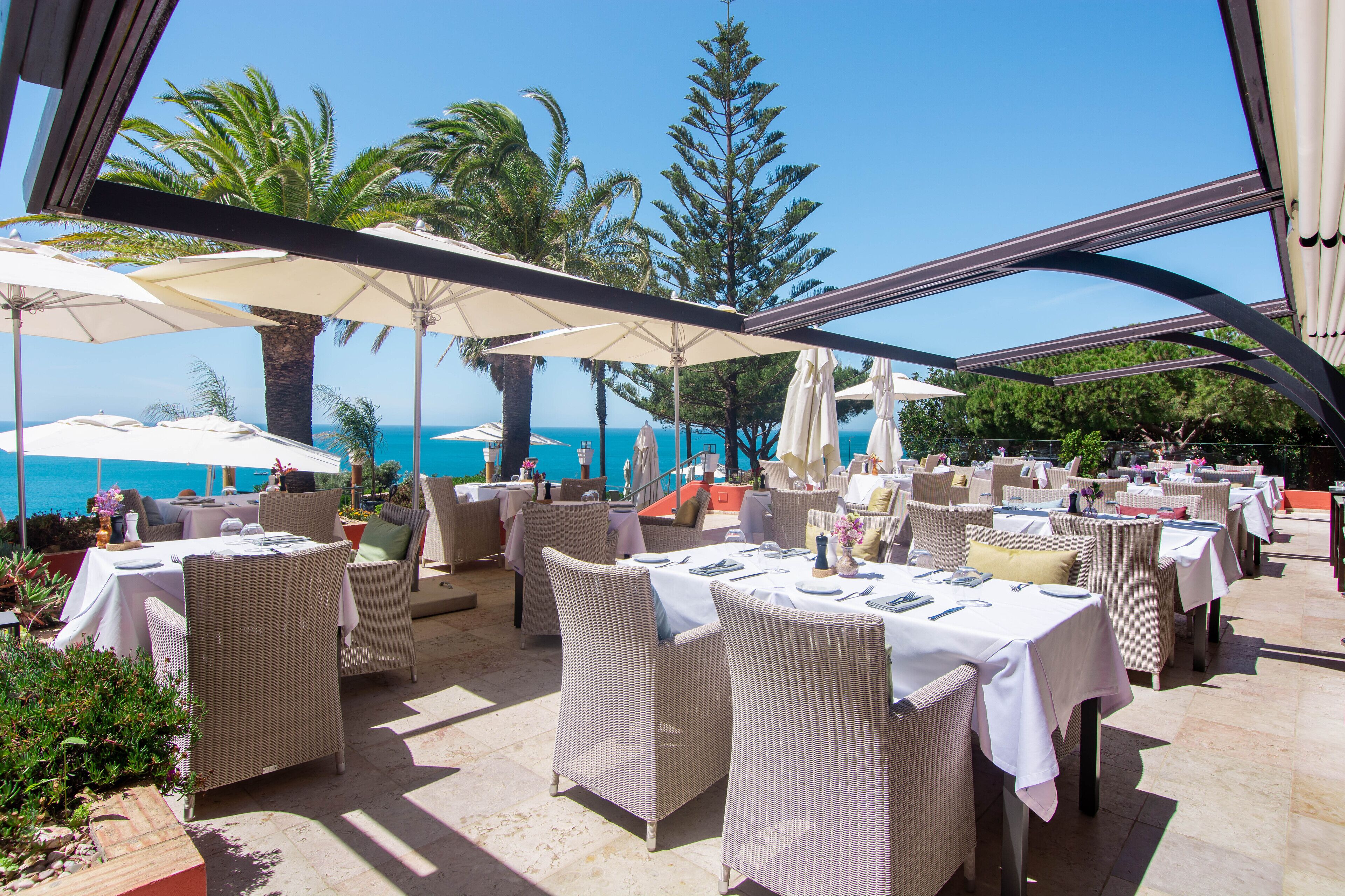 An outdoor dining area with wicker furniture and white tablecloths, overlooking the sea.