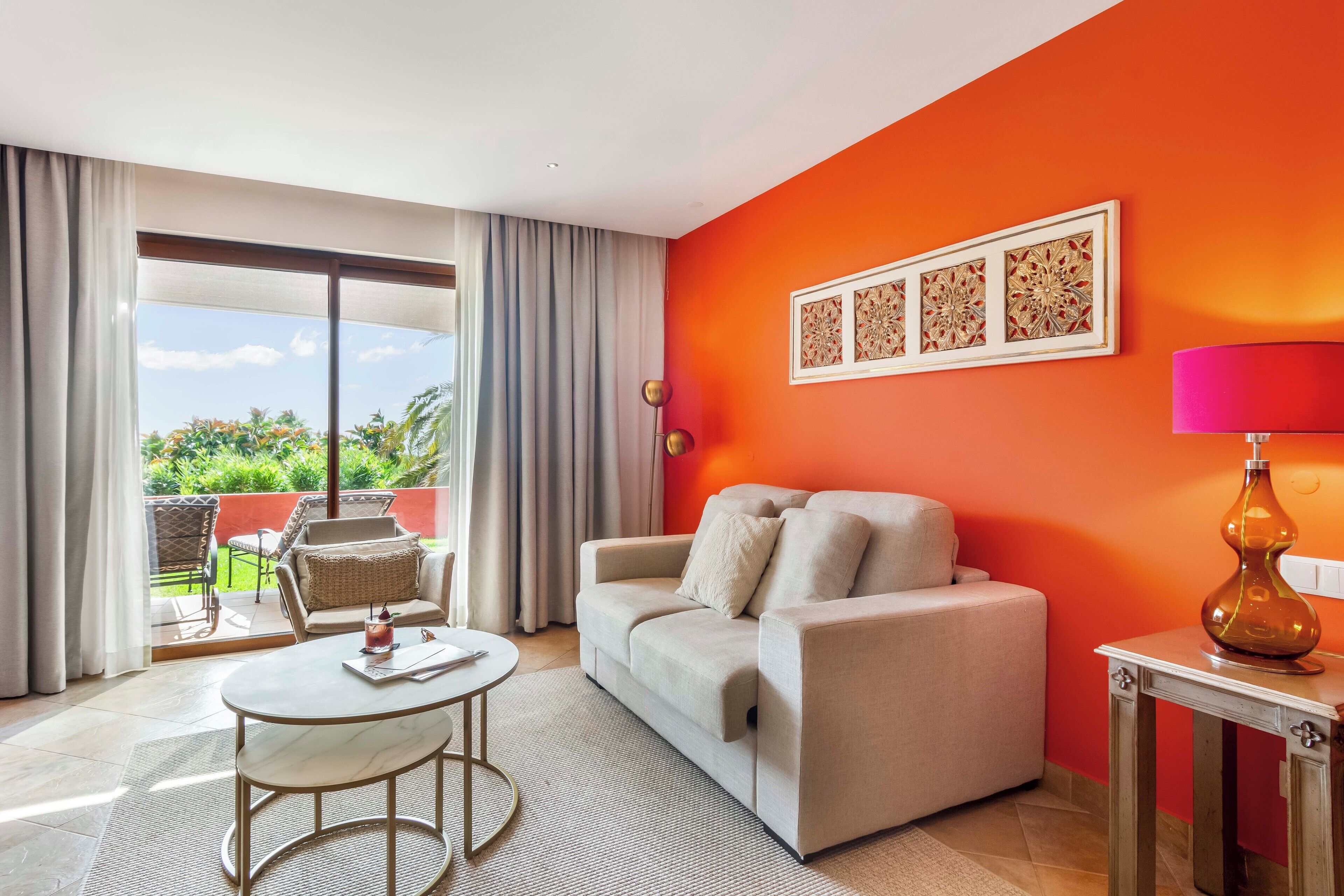 A room with a beige sofa, a round table, and a view of a balcony with outdoor furniture.