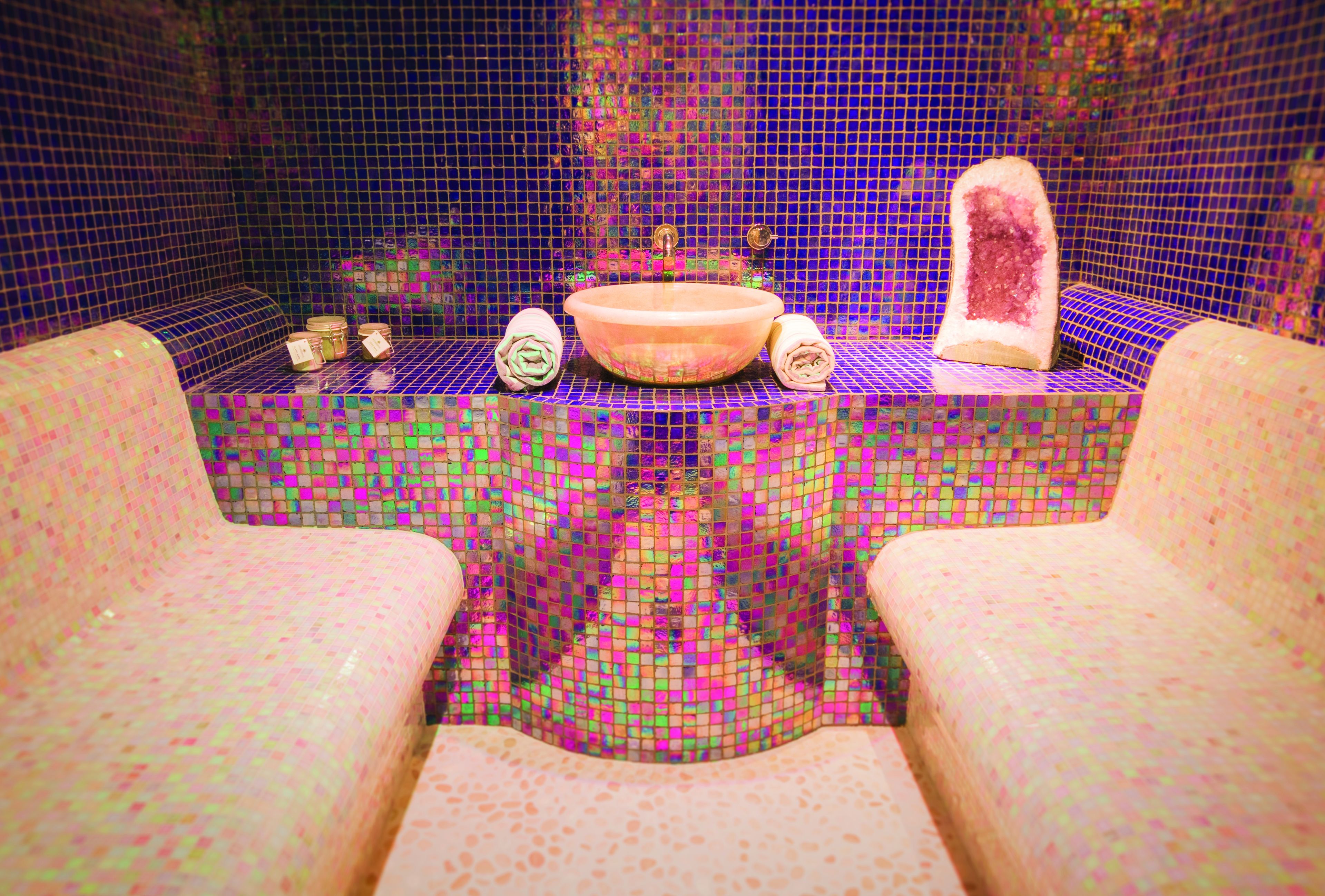 A bathroom with a mosaic tile wall, a central sink, and two towels on a bench.