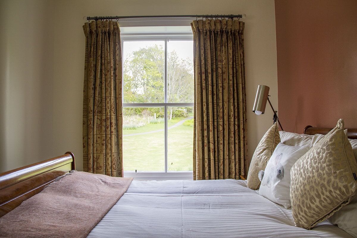 A bedroom with a large window, a bed with patterned pillows, and a bedside lamp.