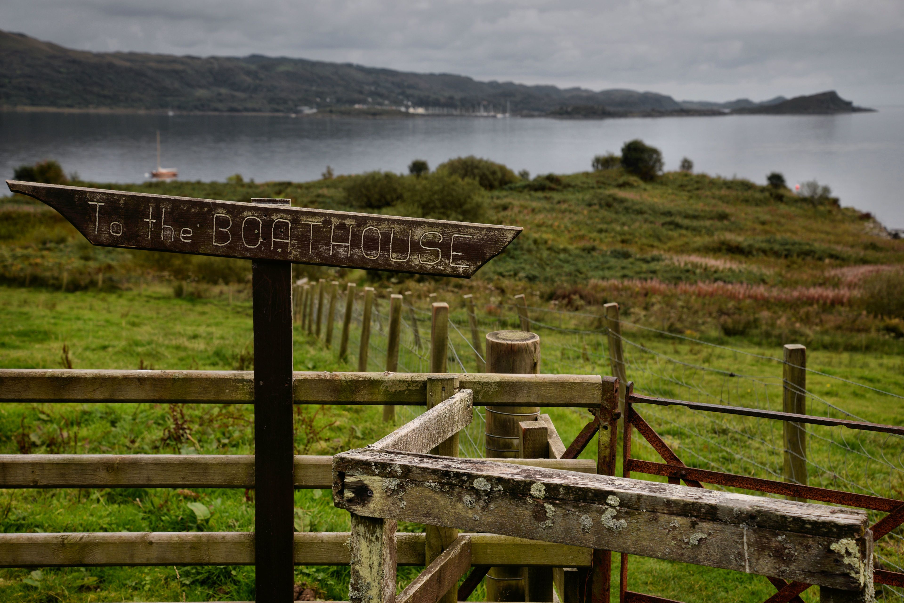 A wooden signpost with the inscription "To the BOATHOUSE" in a rural setting with a body of water and hills in the background.