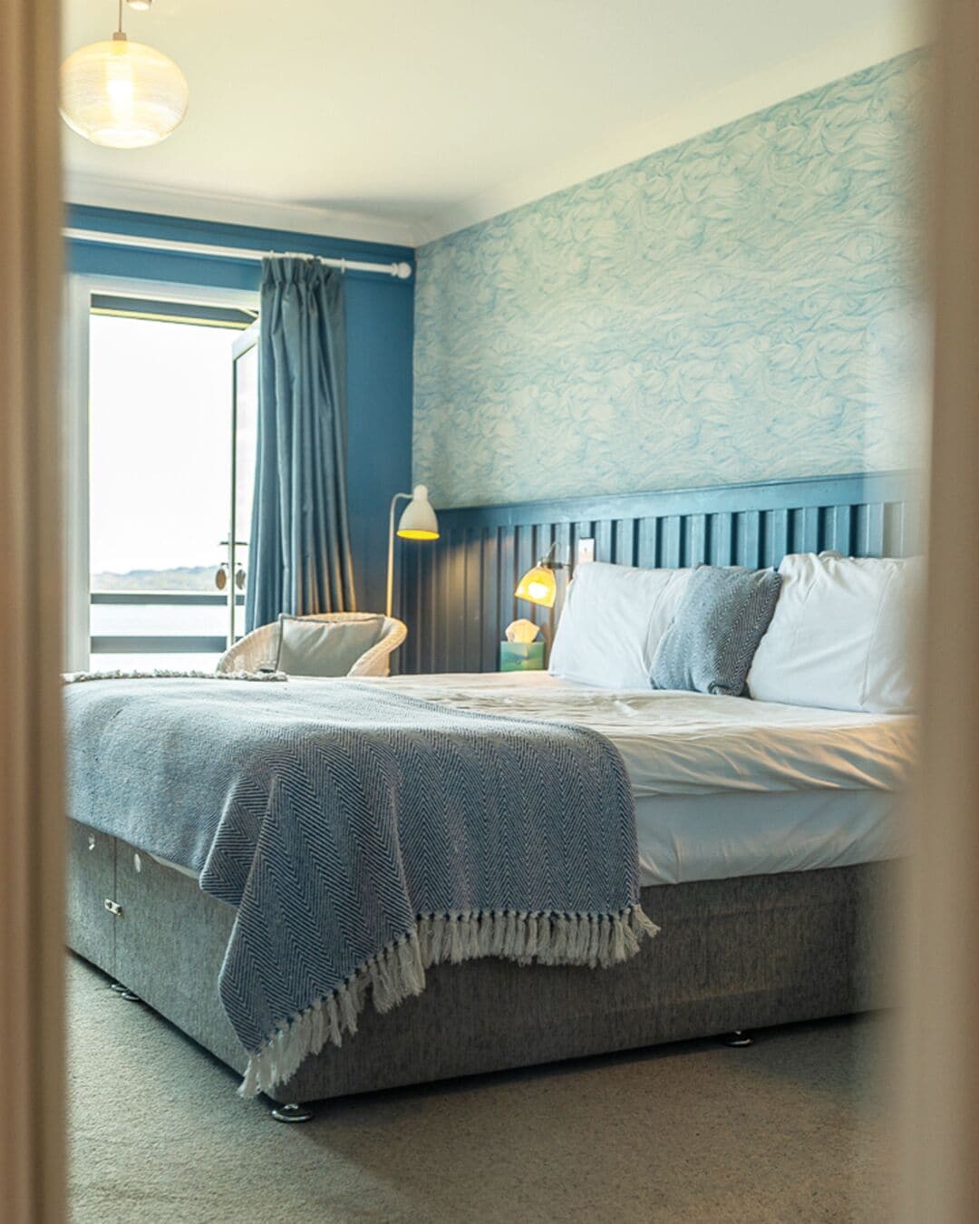 Un dormitorio con cama, una silla, una lámpara y una ventana con cortinas.