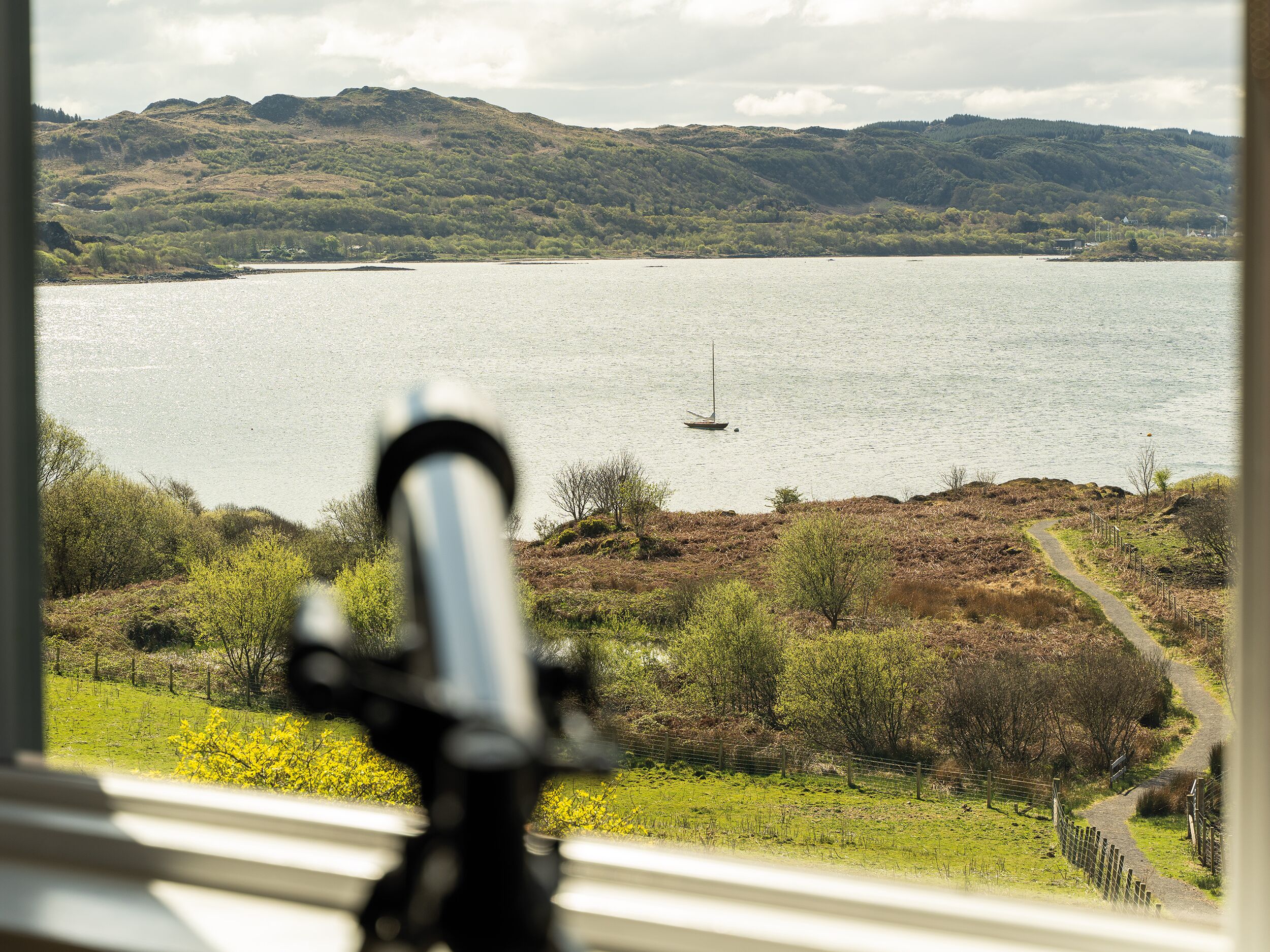 Vista de un lago con un velero, rodeado de colinas y un sendero que lleva a una puerta.