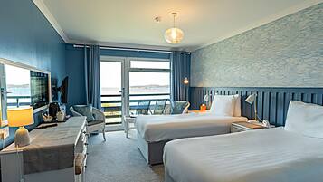 A hotel room with two beds, a desk with a lamp, a chair, a TV, and a view of the sea.