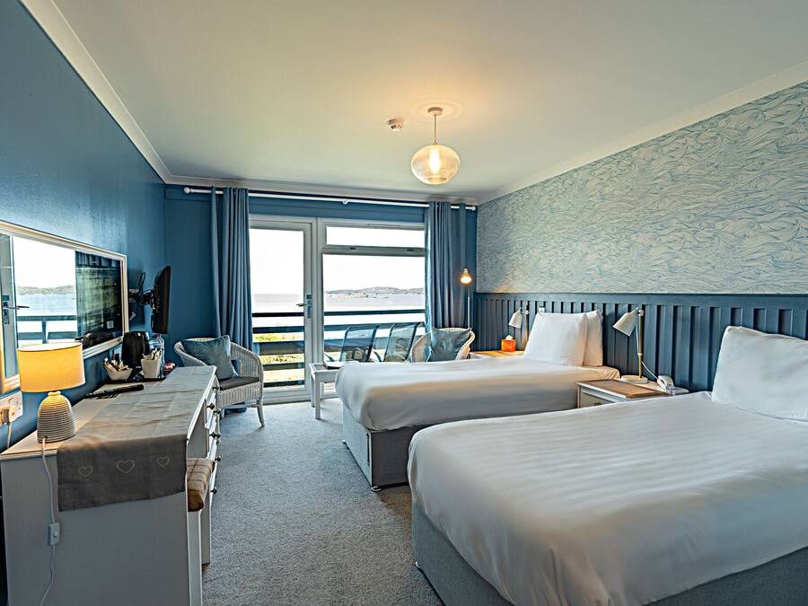 A hotel room with two beds, a desk with a lamp, a chair, a TV, and a view of the sea.
