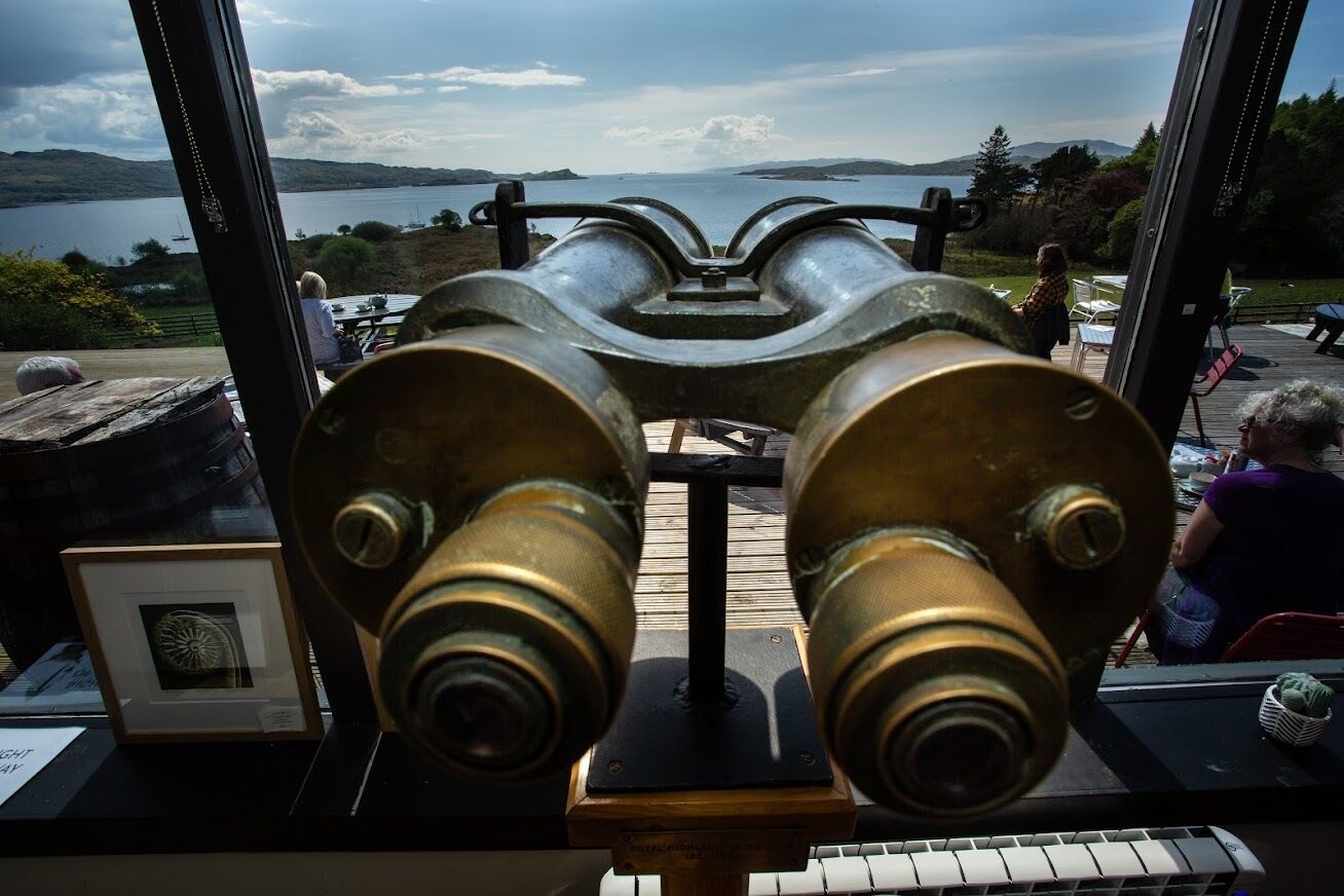 Un télescope en laiton, posé sur un support en bois, offre une vue panoramique sur un lac et des collines, visible à travers une fenêtre.