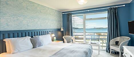 A bedroom with a large bed, a chair, a desk, and a view of the sea.