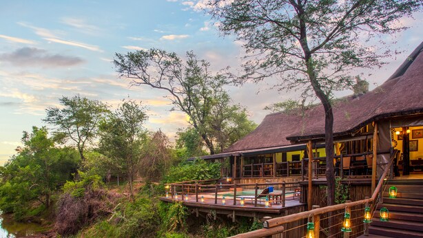 A wooden deck with railings overlooking a river, a thatched-roof building, and a pool area with lanterns. - Madikwe River Lodge (Ramotshere Moiloa)