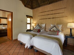 Habitación de hotel con dos camas, cabecera de madera, suelo de baldosas, puerta de madera y ventana con vistas.