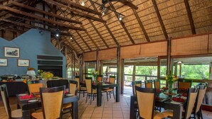 A restaurant with a rustic thatched roof, wooden beams, and tiled flooring. Tables are set with chairs, and there are windows providing natural light. - Madikwe River Lodge (Ramotshere Moiloa)