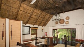A room with a bed, a small table, a chair, a window with a view of greenery, and a ceiling with a thatched roof. - Madikwe River Lodge (Ramotshere Moiloa)