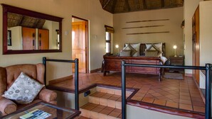 A hotel room with a bed, a sitting area with a chair and a glass table, a wooden dresser, and a ceiling fan. - Madikwe River Lodge (Ramotshere Moiloa)