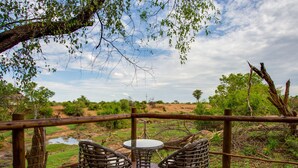 A wooden deck with wicker chairs and a table, overlooking a natural landscape with trees and a river. - Madikwe River Lodge (Ramotshere Moiloa)