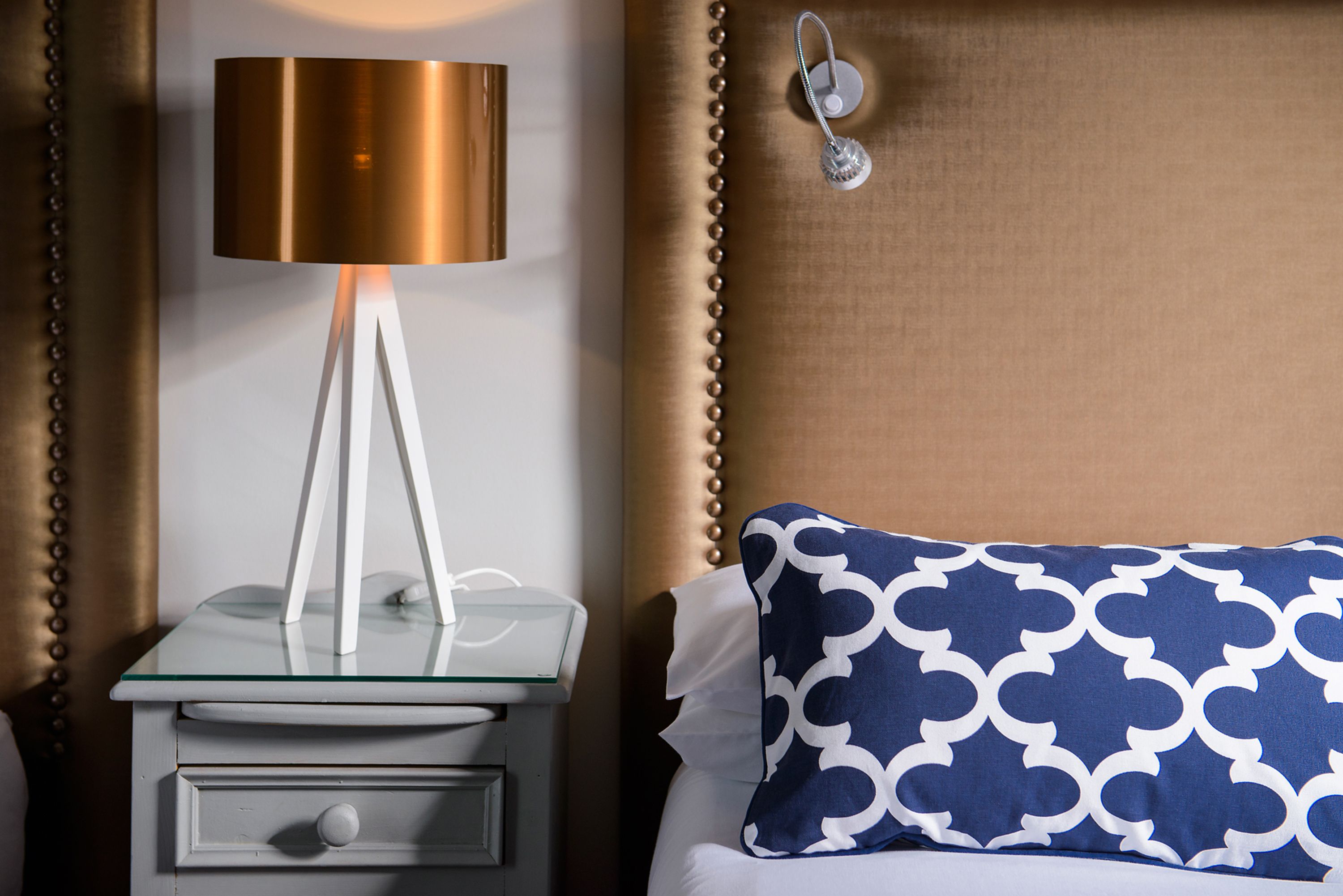 Un lit bien fait, agrémenté d’un coussin à motifs, une table de chevet avec une lampe et un luminaire fixé au mur.