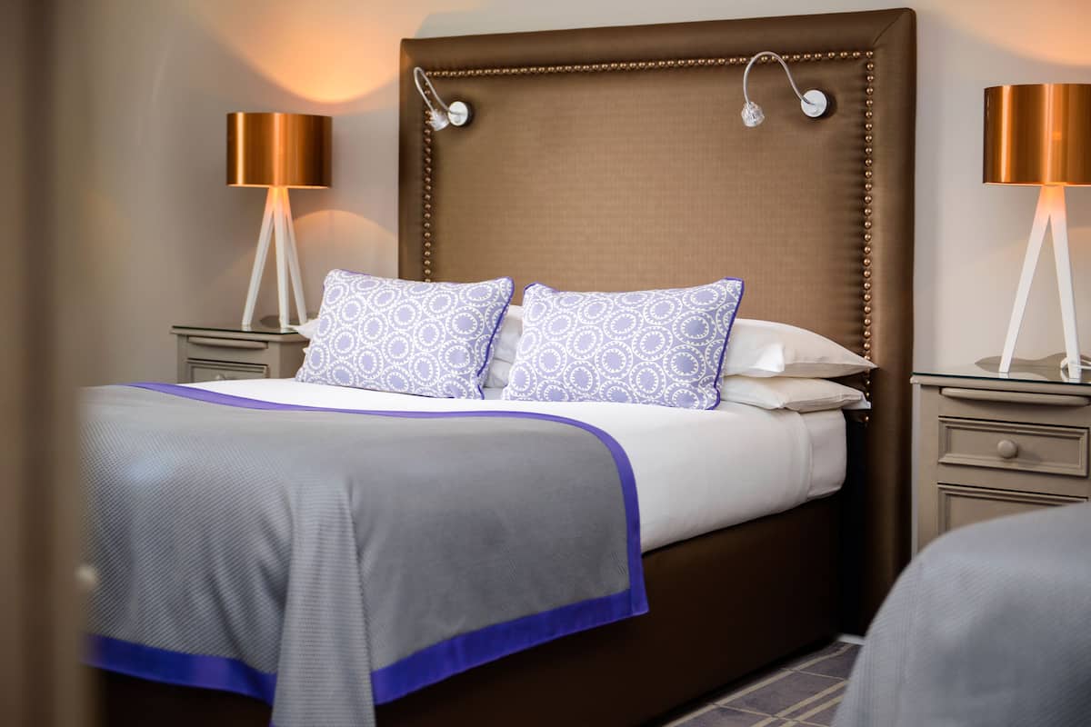 A neatly made bed with patterned pillows, a brown upholstered headboard, and two bedside lamps.
