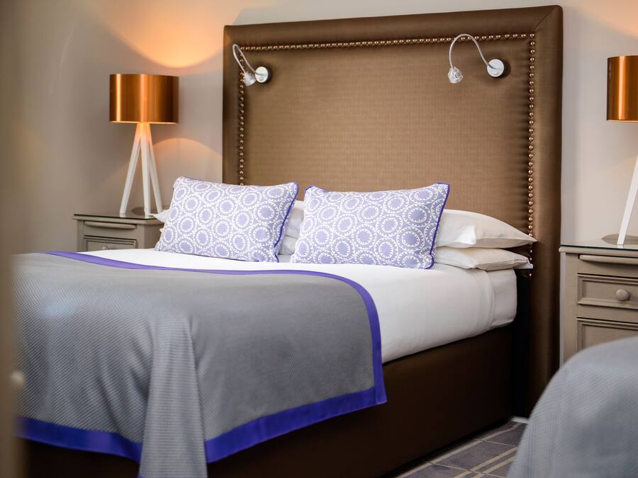 Una cama bien hecha con almohadones estampados, un cabecero tapizado en marrón y dos lámparas de noche.