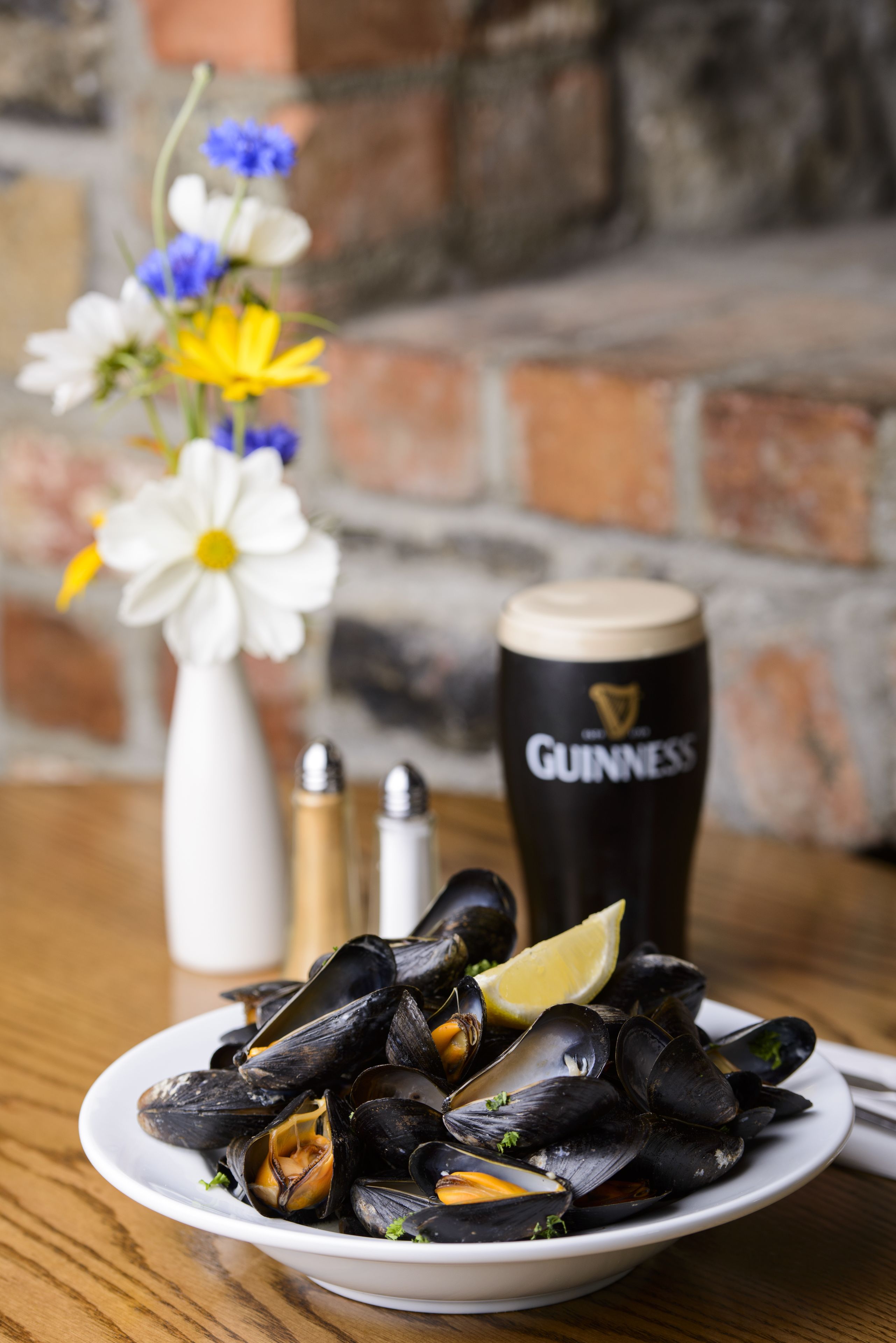 Une assiette de moules accompagnée de quartiers de citron, un vase blanc rempli de fleurs et un grand verre de bière Guinness sur une table en bois.