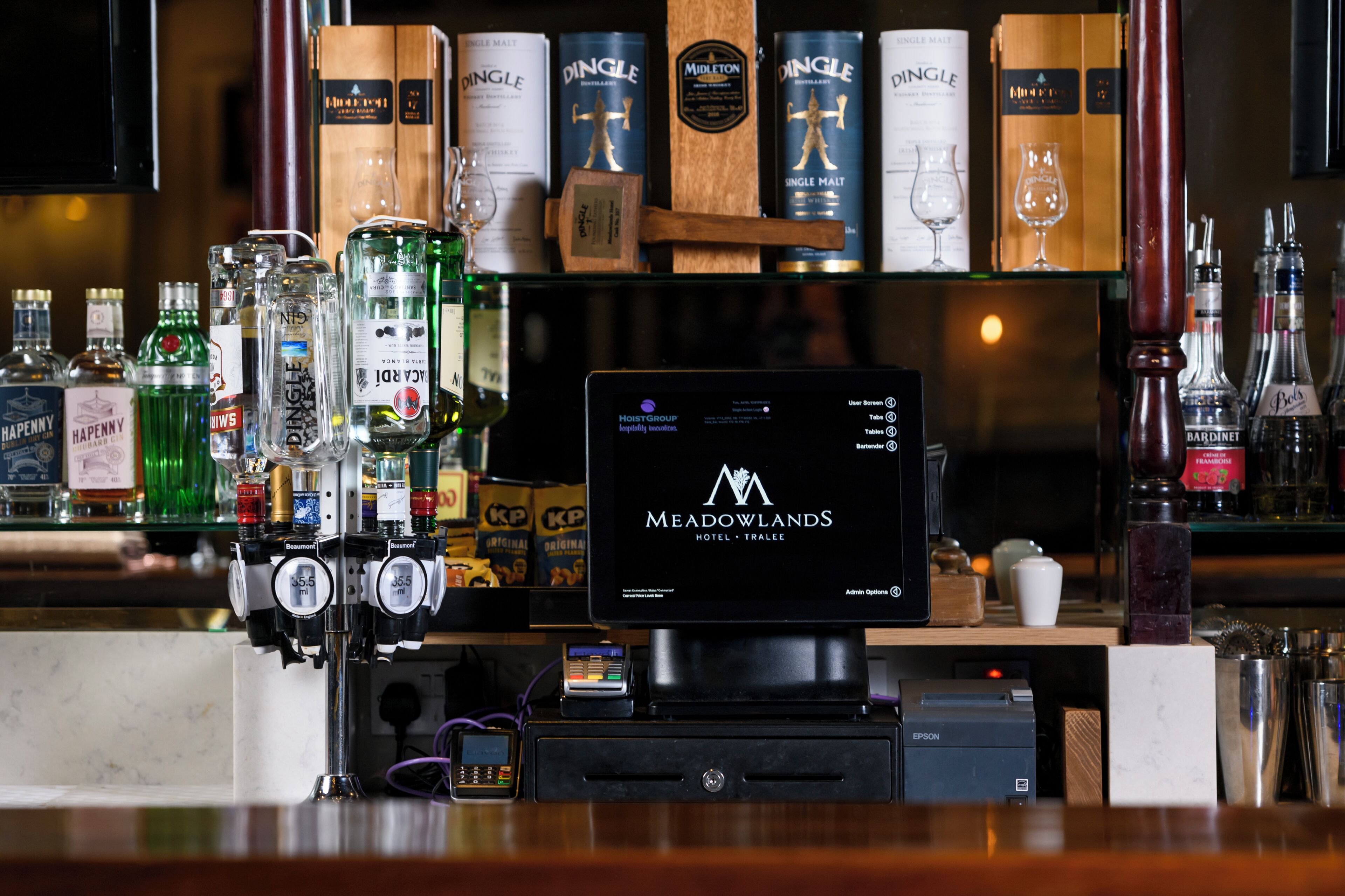 Zona de bar con una pantalla digital que muestra 'MEADOWLANDS HOTEL - TRALEE', varias botellas de licor y una caja registradora.