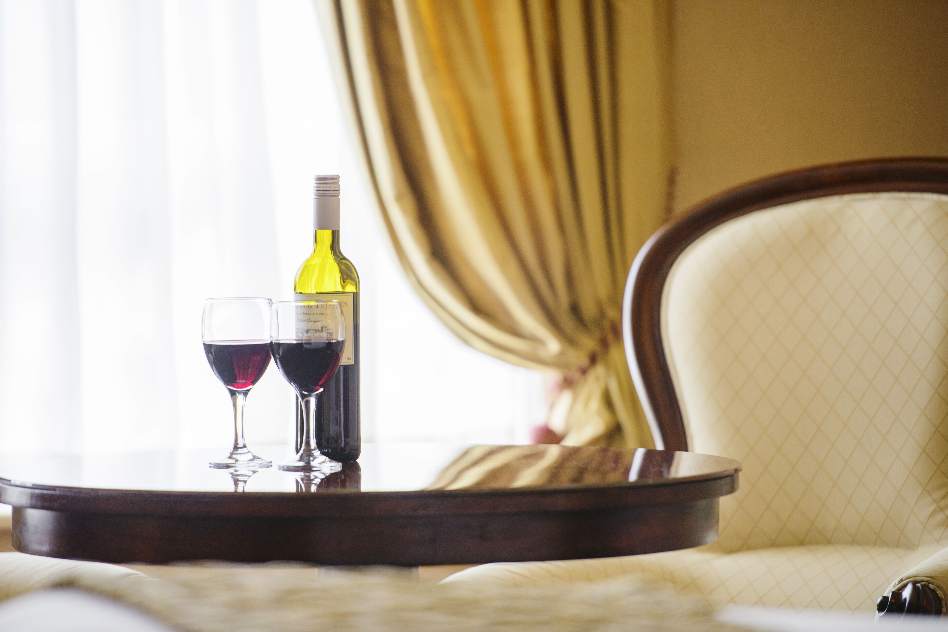 Een tafel met een fles wijn en twee glazen, in een kamer met een stoel en gordijnen.