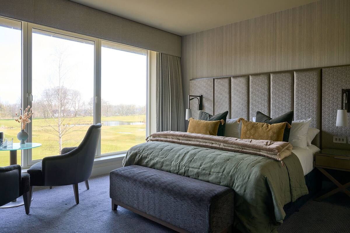 A bedroom with a large window, a bed with multiple pillows, a bedside table, and a chair.