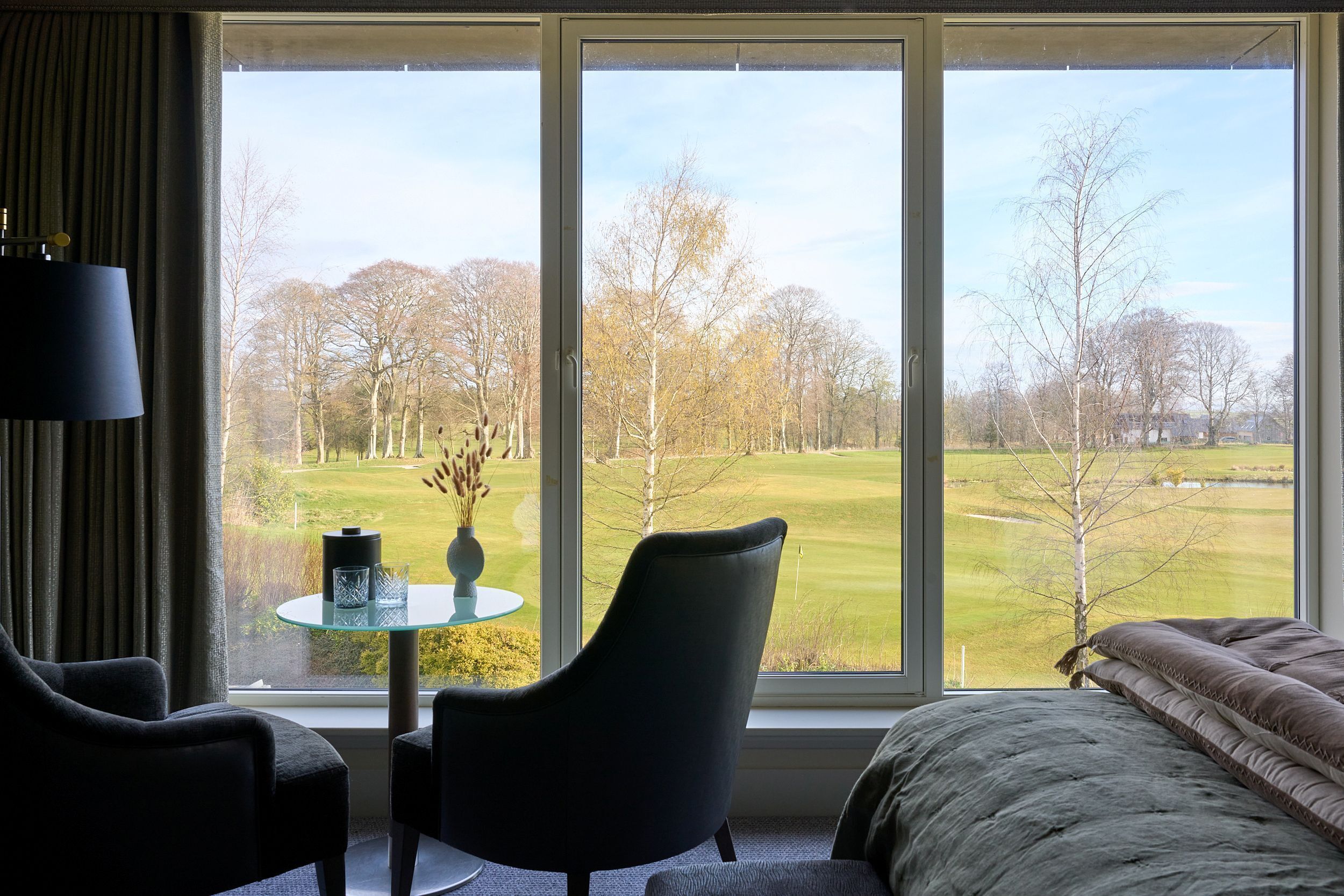 Une pièce avec une grande fenêtre, un lit, une chaise et une petite table sur laquelle se trouvent un vase et des verres.