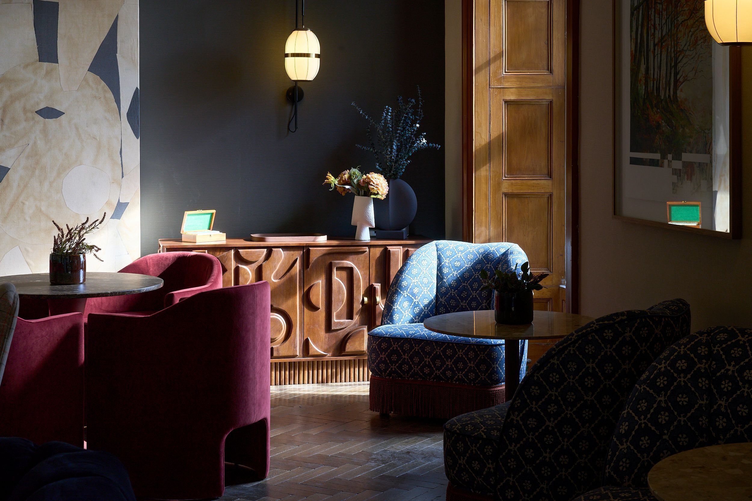 A room with a wooden cabinet, a blue patterned sofa, a red chair, a small table with a plant, and a wall-mounted light fixture.