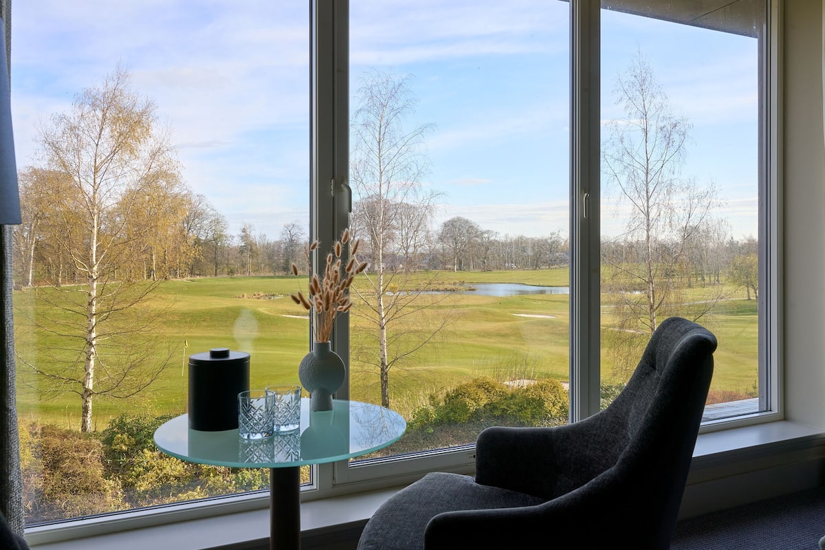 A room with a large window, a round glass table with a vase and a black cylindrical object, and a grey chair.