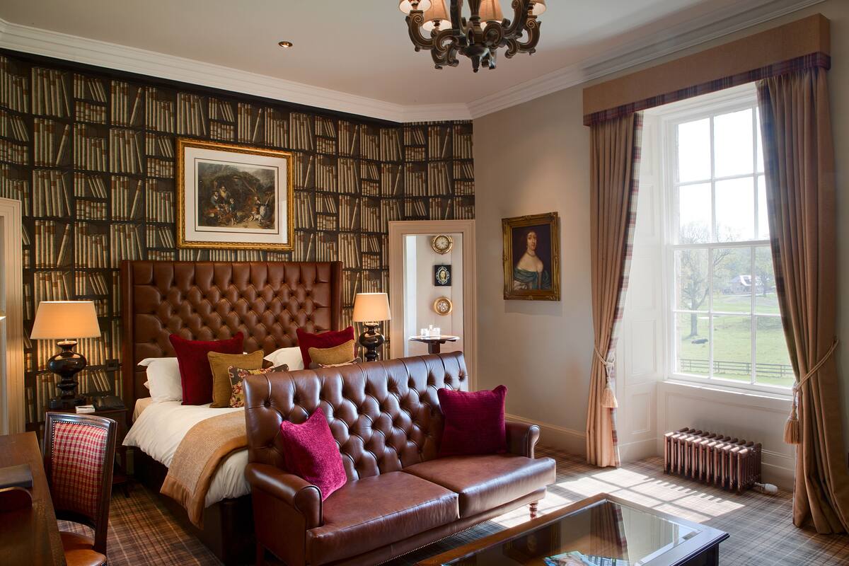 A room with a tufted leather sofa, a large bed with multiple pillows, a bookshelf, a painting, a mirror, and a view of the outdoors through a window.