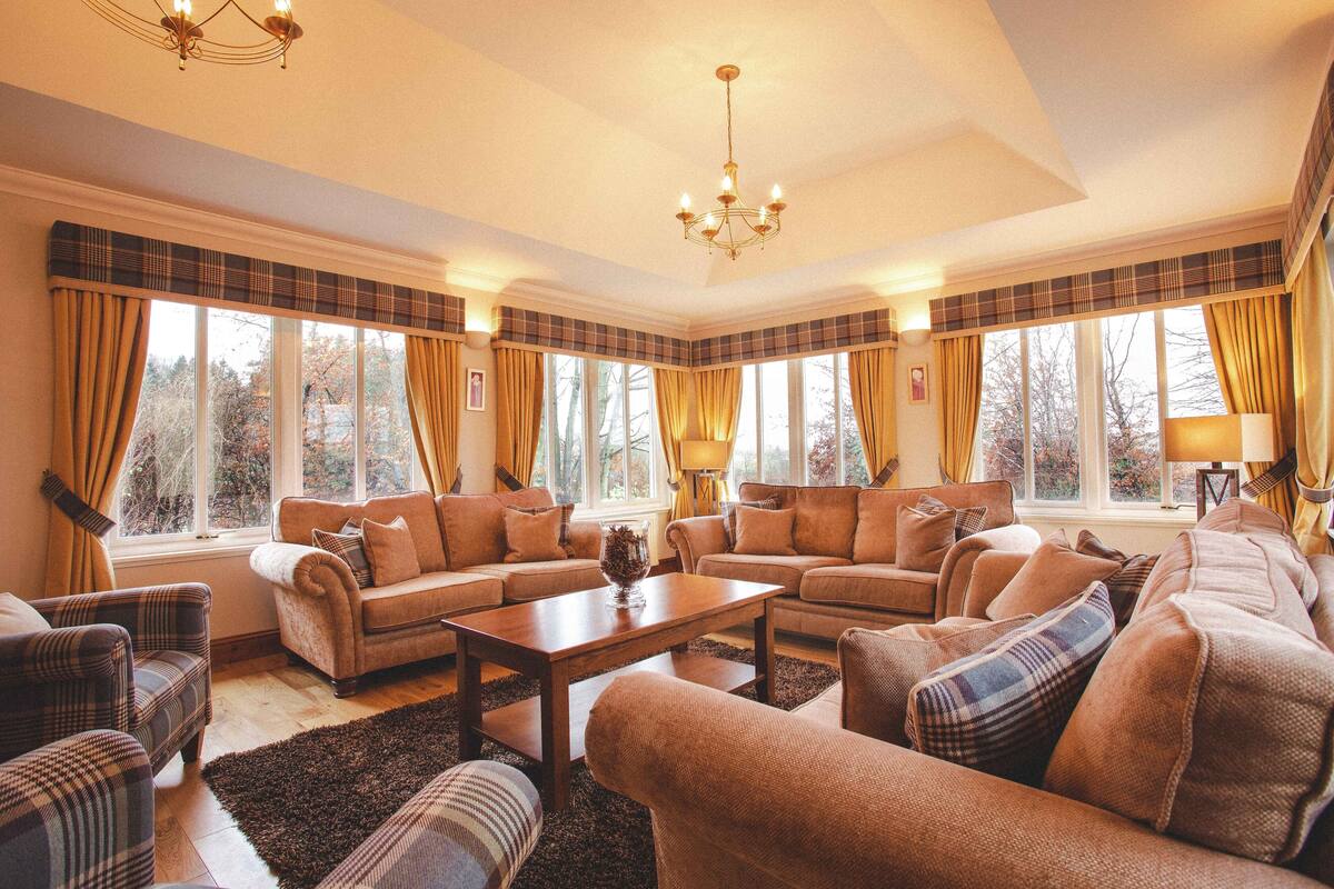 A spacious living room with a fireplace, large windows with curtains, and a chandelier.