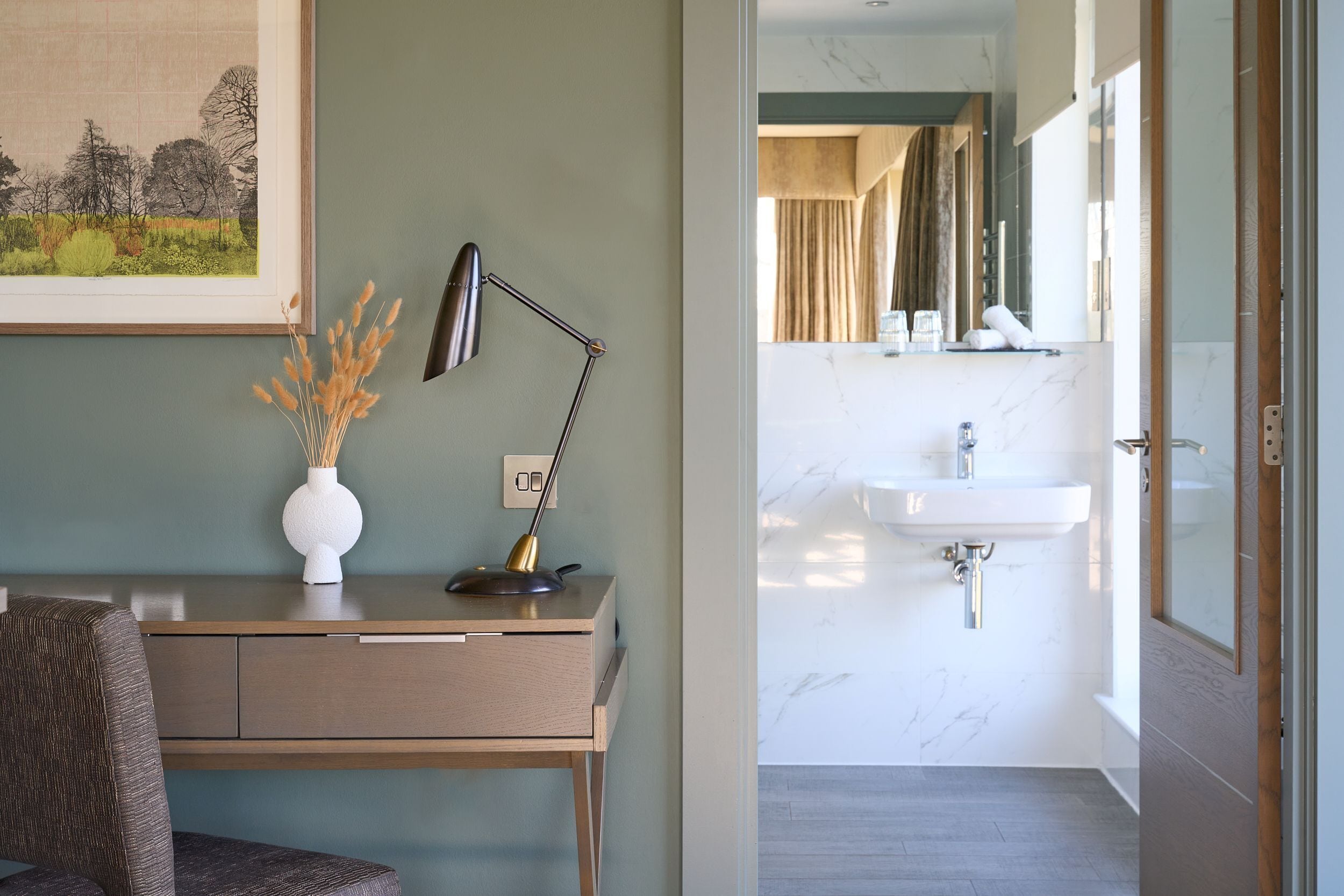 A modern room with a desk, a lamp, a vase with dried plants, and a view of a bathroom with a sink and mirror.