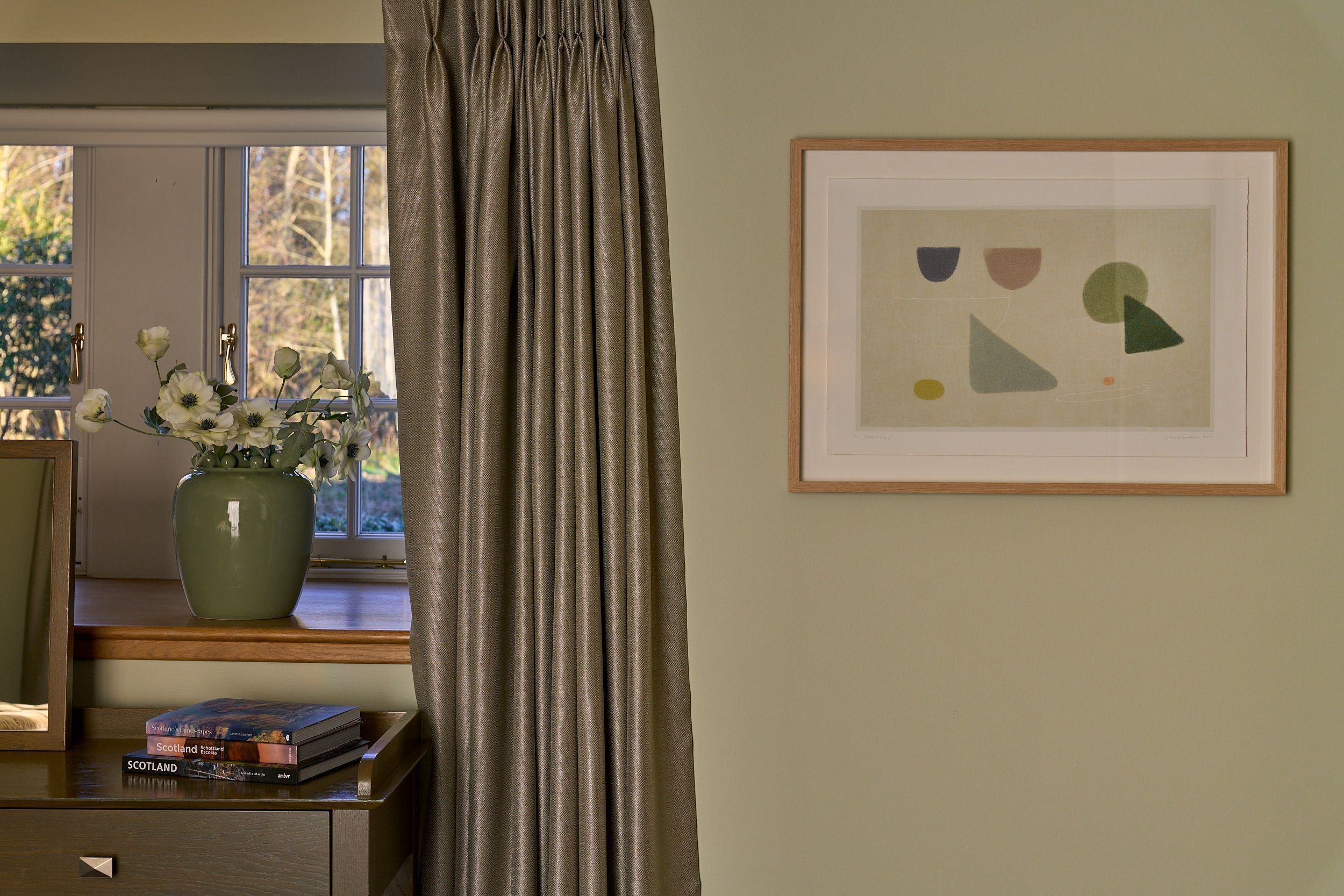 A room with a window, a framed abstract artwork, a vase with flowers, and a stack of books on a desk.